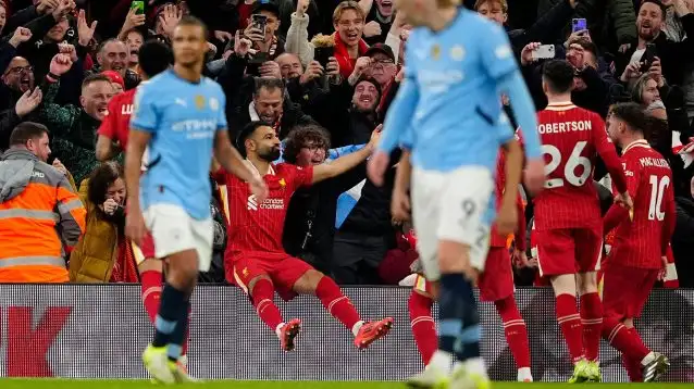Mo Salah sits on the advertising hoarding with celebrating fans and teammates around him as Man City players trudge away in the foreground