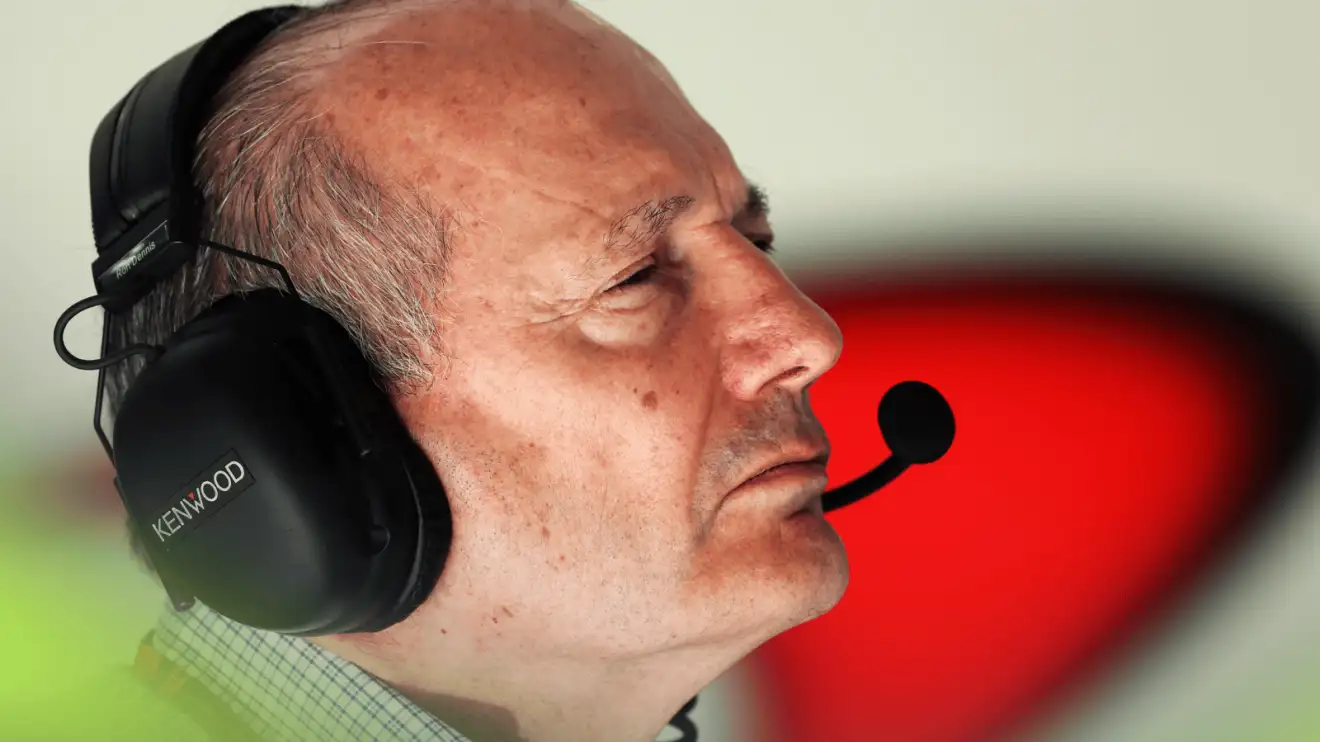 A close-up shot of Ron Dennis wearing a headset in the McLaren garage with the team's 'speedmark' logo in the background